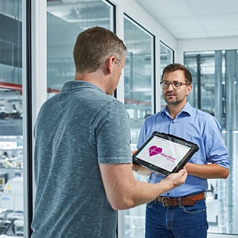 Ein grauhaariger mann spricht mit einem dunkelhaarigen mann. Der Grauhaarige hält ein Tablet in der Hand